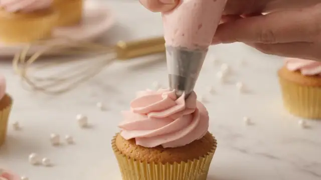 Hands piping pink buttercream onto a cupcake